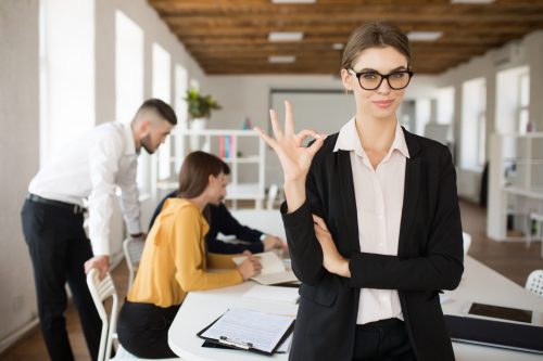 Young smiling business woman in eyeglasses and shirt dreamily looking in camera showing ok gesture spending time in office with colleagues on background